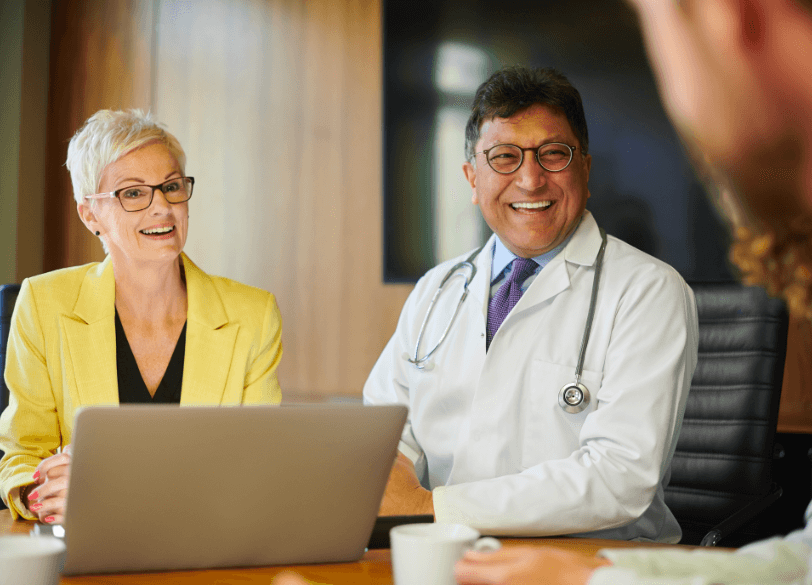 A physician in a white coat and a colleague in a yellow blazer smile during a meeting at a laptop.