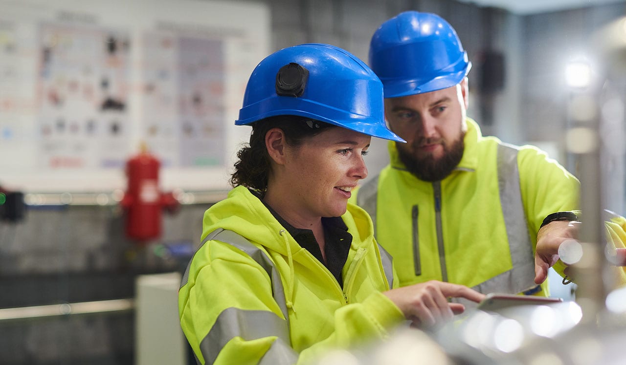 To ansatte med hjelm og refleksvest ser på et nettbrett i en fabrikkhall